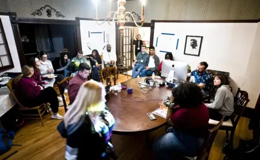 Students sit around a table with computers and notebooks in lively discussion.