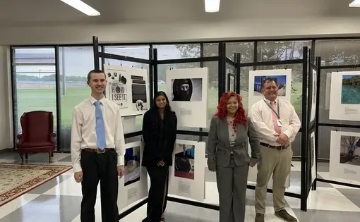 Three students and their teacher pose in front of the display of images from the How I See It Photography Exhibition
