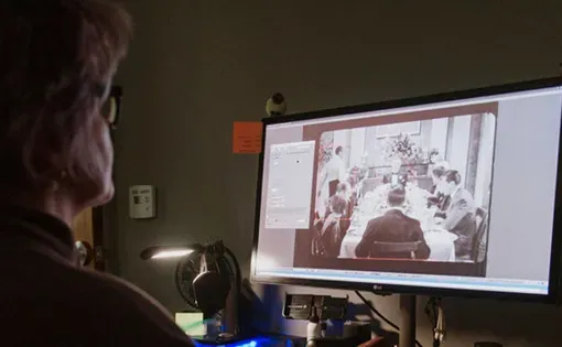 A woman monitors a computer screen that shows an image of a meeting of men in suits in a formal setting. There are red squares surrounding different figures in the image.