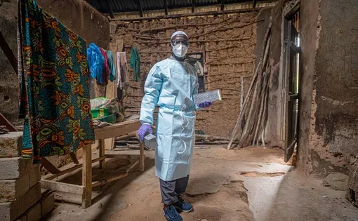 A medical professional in protective wear, gloves, mask and goggles holds two metal boxes as he stands in the middle of an building made from mud bricks