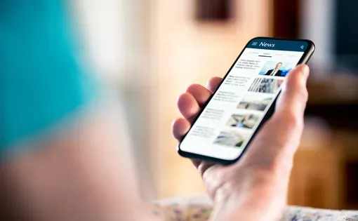 An adult holds a smart phone in their hand where news headlines are visible on the screen.