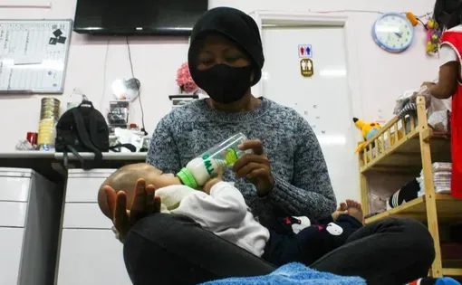 A woman wearing a mask and head covering feeds a baby with a bottle.
