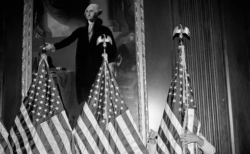 A person stands behind American flags next to a portrait of George Washington