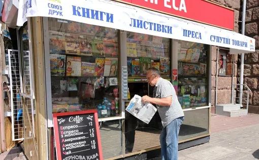 A man in a white shirt and blue jeans looks at a newspaper at a newspaper stand.