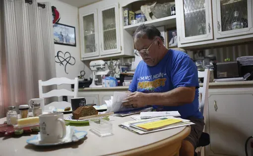 A man wearing a blue shirt sits at a table looking through papers in his home