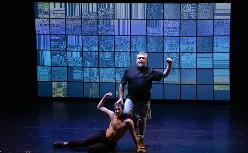 On a well lit stage, one man kneels at the feet of another, who raises a fist into the air.