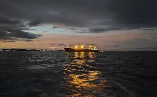 A cargo ship at sea at dusk, under a gray sky. Yellow lights from the ship reflect on the water, creating a dramatic and atmospheric maritime scene. Ideal for maritime, shipping, and travel themes
