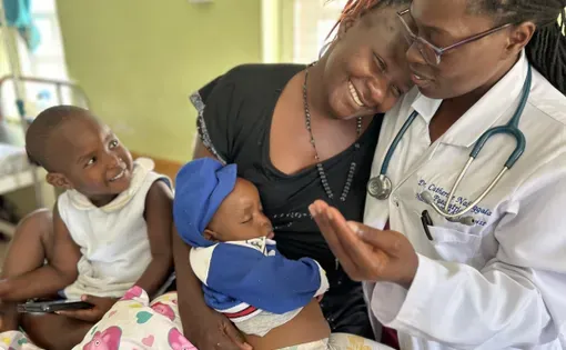 A toddler wearing a white sleeveless shirt smiles while watching a smiling woman hold a baby and laying her head on a doctor's shoulder.