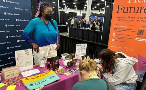 A person stands behind a table at an exhibit booth, talking to students who are seated and writing.