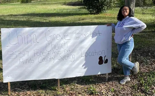 Larnee Satchell poses at the empty field, where Mississippi’s Statewide Election Management System erroneously directed voters, posing with a sign directing them to the proper location. Hinds County election officials placed the sign there after she contacted them about the issue on March 12, 2024. Photo courtesy Larnee Satchell