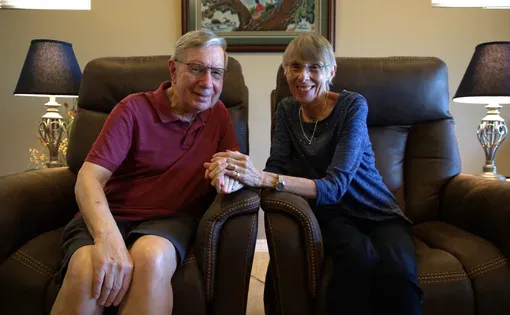 A smiling, elderly couple hold hands while sitting down and facing the camera.