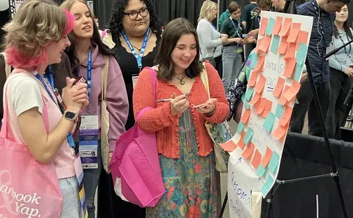 Three students and a teacher talk, laugh, and fill out post-its to stick on a posterboard in a conference exhibit hall.
