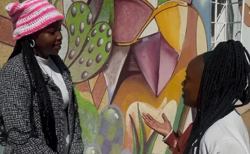 Two young women talk in front of a colorful mural.