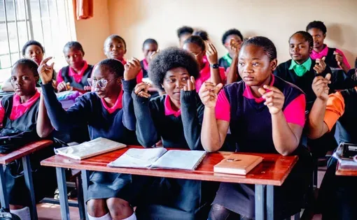 Female students sit in a classroom at their desks, snapping their fingers.