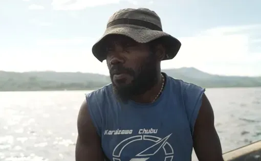 A fisherman wearing a light blue sleeveless shirt and tan hat sits on a small boat