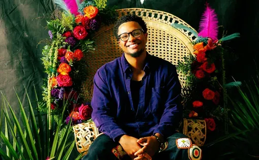Adam Mahoney sits in a rattan chair decorated with tropical flowers and feathers. Neon sign behind him says "beaucoup power: Black is brilliant". Indeed it is.