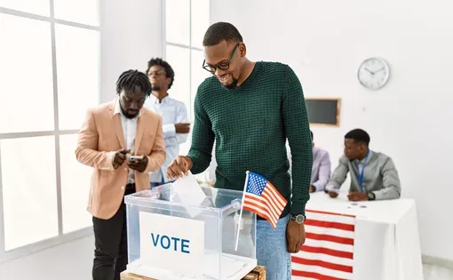 A man wearing a green sweater places a ballot in a box labeled "Vote."