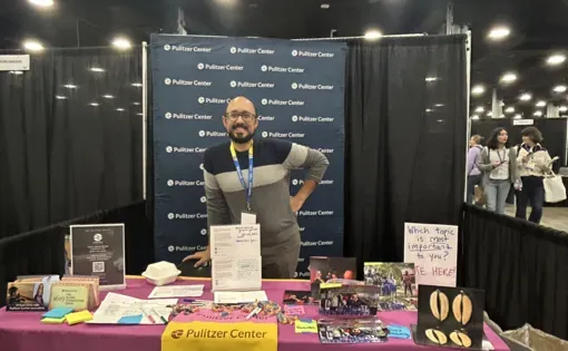 Fareed stands in a conference booth behind a table decorated with Pulitzer Center tablecloth and several Pulitzer Center-themed activities and materials.