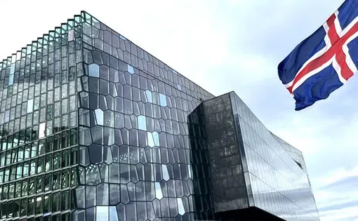 The Icelandic national flag waves outside a large building covered entirely with organically-shaped glass panels.