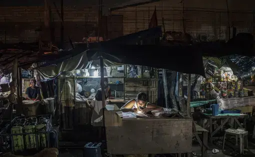 A woman works on bookkeeping using the light of her phone in the Market 3 slum in the Navotas Fish Port Complex. Market 3 has electricity but no plumbing. Residents often turn to drugs to alleviate hunger and stay awake for the long hours they work at low pay. Image by James Whitlow Delano. Philippines, 2017.