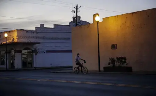 A man riding a bicycle down an empty street wears a protective mask amid the COVID-19 coronavirus outbreak on Friday, April 17, 2020, in Dawson, Ga. Image by Brynn Anderson / AP Photo. United States, 2020.