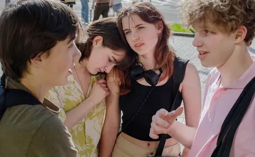 Four teenagers stand together, gathered in conversation on a sunny day. One girl leans on the other's shoulder.