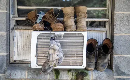 Boots and a hat dry on a window air conditioning unit at a Johnston County farmworker camp Thursday August 27, 2020. Air conditioning is considered a luxury by many farmworkers in North Carolina farmworker camps. Image by Travis Long. United States, 2020.