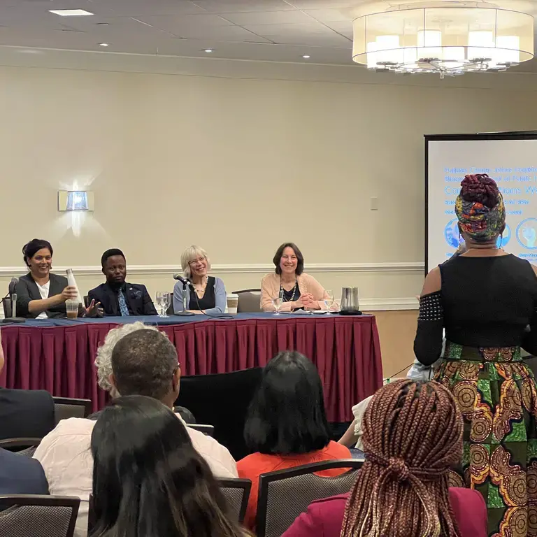 A line of speakers sit at a panel table in front of a room filled with an audience. One person is standing up in the audience facing the panel.