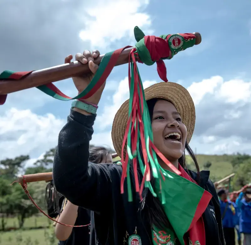 A young woman holds up a stick decorated with green and red ceremonial ribbons. She stands in a crowd of other people also raising similar sticks.