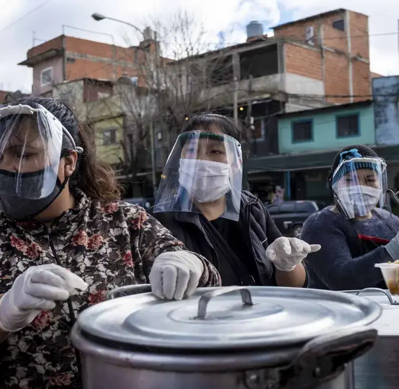 a line of women wearing protective gear and masks stand behind pots to hand out