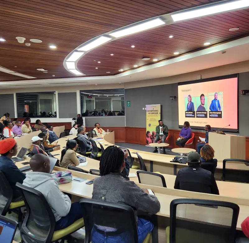 a conference room filled with rows of tables with a group of people listening to someone in the front