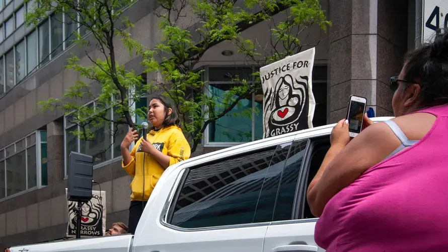 Jenae Turtle (left) stands before a crowd and speaks about her community while Chrissy Isaacs (right) stands below her, filming Jenae's speech. Canada, 2019. Image by Shelby Gilson.
