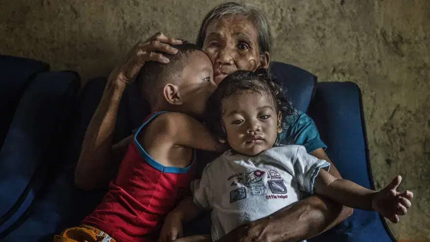 Remy Fernandez, 84 years old, holds two of the seven grandchildren she is raising. Her youngest son, Constantino de Juan, a methamphetamine user, was killed by masked men in December, 2016. Upon seeing his attackers, Juan instructed five-year-old CJ, shown here wearing a red tank top, to take care of his siblings. The children's mother is in prison due to a drug arrest. Baby RJ, in the 'daddy's little helper' T-shirt, was born in prison. Image by James Whitlow Delano. Philippines, 2017. 