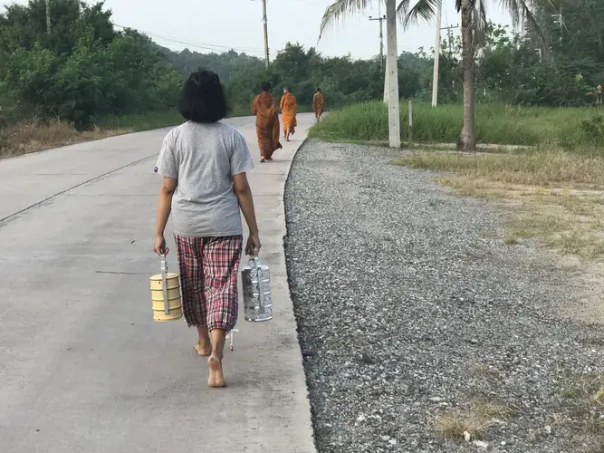 A young Thai girl follows three novice monks in the collection of the morning alms, during which they accept donations of food and drink to the temple from residents throughout the village of Chonburi. Image by Kiley Price. Thailand, 2018.