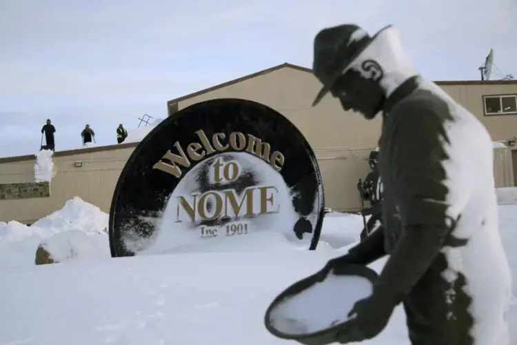 In this Feb. 14, 2019, photo, three men shovel snow from the roof of a grocery store as a statue of one of the 'Three Lucky Swedes,' credited with discovering gold in the late 19th century, stands in the foreground, in Nome, Alaska. The city later added statues of two native boys who led him to find the gold. Image by Wong Maye-E. United States, 2019.