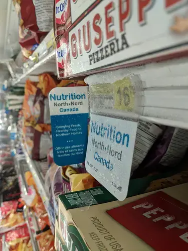 At a store in Pangnirtung, Nunavut, a sign for the Nutrition North program hangs above a frozen pizza (cost: $16.79). Inuit groups have been pressing Ottawa to reform the controversial subsidies for southern food flown into the North. Image by Julie De Meulemeester. Canada, 2018.