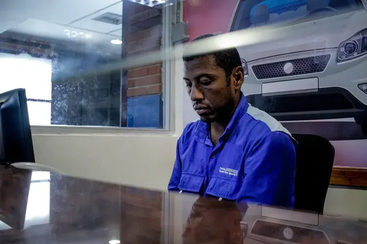 Carlos Román sits at his job post, his eyes staring down, wearing a bright royal blue uniform shirt. He works as a cashier in a parking lot at a shopping mall. He works the afternoon shift, from 2 p.m. to 7 p.m. It usually takes several hours to get back home to the satellite city of Guarenas because of the lack of public transportation. Image by Flaviana Sandoval. Venezuela, 2018.