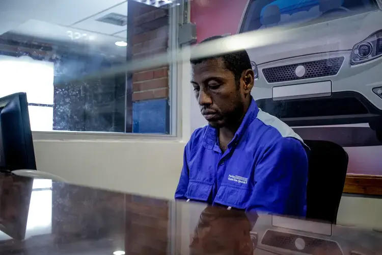 Carlos Román sits at his job post, his eyes staring down, wearing a bright royal blue uniform shirt. He works as a cashier in a parking lot at a shopping mall. He works the afternoon shift, from 2 p.m. to 7 p.m. It usually takes several hours to get back home to the satellite city of Guarenas because of the lack of public transportation. Image by Flaviana Sandoval. Venezuela, 2018.