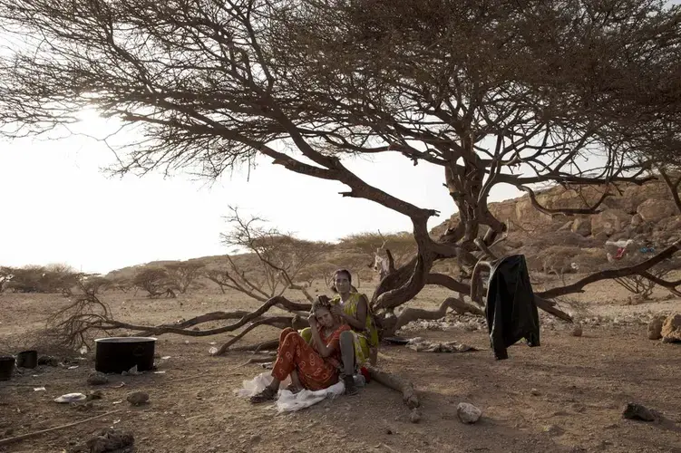 Fatma, right, a migrant from Ethiopia, braids her friend's hair, as they take shelter on the last stop of their journey before leaving by boat to Yemen in the evening, in Obock, Djibouti. Image by AP Photo/Nariman El-Mofty. Djibouti, 2020.