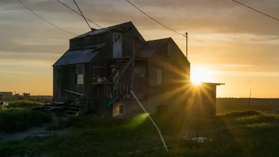 Rays of sunlight spill over a Shishmaref home as the midnight sun sets over the Chuckchi Sea. Image courtesy of Nick Mott. United States, 2018.