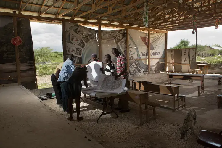 From left: Estimei Volmy, Simeus Salma, Louis Midelove, Pierrelien Patrick, Victor Layers and Condiac Julien look at maps provided by UNA/Habitat of developments that are slated for their community at a meeting of the Table Quartier d’Onaville community group, in a church building made of wood and vinyl posters, in the Onaville neighborhood of Canaan. Image by Allison Shelley. Haiti, 2018.