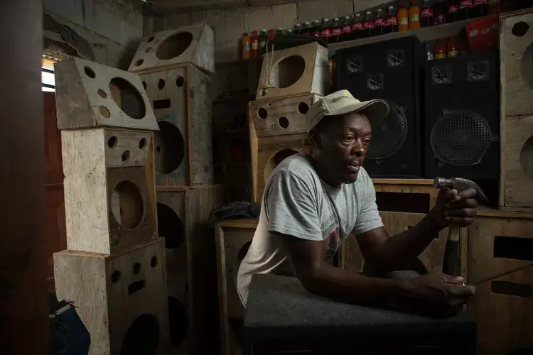 A vendor stands in a neighborhood snack shop (his wife’s business) and a workshop for making speakers (his business) in Canaan’s Corail neighborhood. He makes the speakers by hand from plywood, felt and parts salvaged from broken speakers that he buys secondhand. The biggest units he sells to discos and DJs for over $600 (U.S.) apiece. Lately, business has been slow and he has decided to sell the shop so he can afford to buy the parts to finish a few more, which he plans to set up to play music onto the street to attract customers. Image by Allison Shelley. Haiti, 2018.