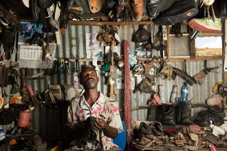 Eddy Bien Aime repairs a pair of sandals at his business on the main road to the Canaan II section of Canaan. Most of Bien Aime’s business involves selling footwear that he has bought secondhand and refashioned. He moved to this area on Jan. 16, 2010, just days after the quake, and took over a large parcel, some of which he has since given away. “Downtown, there are killings. I have to be careful when I come back from there that people don’t rob me. Here in Canaan, it feels better,” says Bien Aime. Image by Allison Shelley. Haiti, 2018.</p>
<p>