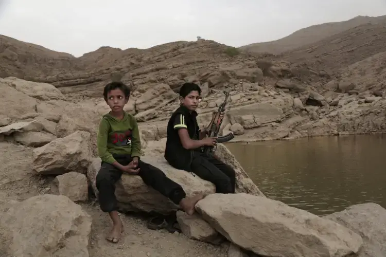 In this July 30, 2018 photo, a 17-year-old boy holds his weapon in High dam in Marib, Yemen. Experts say child soldiers are 'the firewood' in the inferno of Yemen's civil war, trained to fight, kill, and die on the front lines. Though both sides in the war recruit children, the Houthi rebels rely on them the most. One Houthi military official said 18,000 children had been recruited over the past four years. Image by Nariman El-Mofty / AP News. Yemen, 2018.