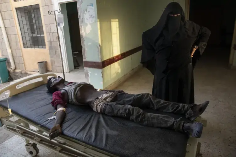 An Ethiopian Tigray migrant who was imprisoned by traffickers for months, lies on a gurney accompanied by a nurse at the Ras al-Ara Hospital. Nurses gave him fluids but he died several hours later. Image by Nariman El-Mofty. Yemen, 2019.