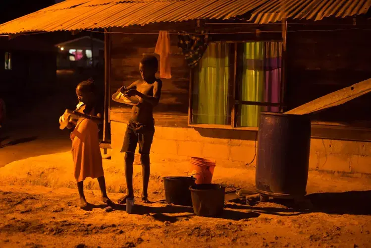 Children clean their shoes for school in the orange glow of a street light on Wednesday, March 15, 2017 in the village of Adjuma Kondre, Suriname. It will take Alcoa years to repair thousands of acres of mined land around the village. Image by Stephanie Strasburg.<br />