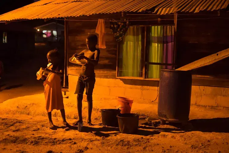 Children clean their shoes for school in the orange glow of a street light on Wednesday, March 15, 2017 in the village of Adjuma Kondre, Suriname. It will take Alcoa years to repair thousands of acres of mined land around the village. Image by Stephanie Strasburg.<br />
