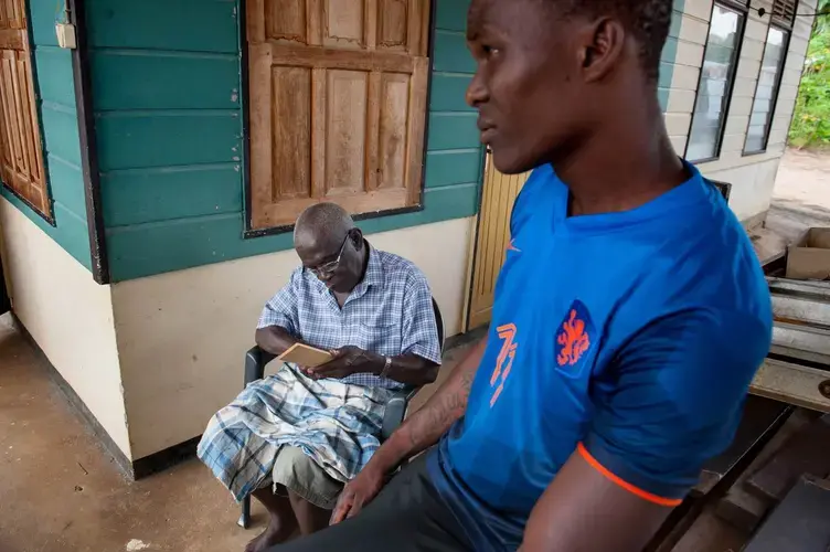 Hiskia Jabini, left, 81, demonstrates making wood combs beside Gieluen Jabini, 32, on his porch in the village of Nieuwe Aurora, Suriname. The elder Jabini makes the combs to sell to the slow trickle of tourists that come through the village, to supplement his government check and the $27 a month pension from years working as a laborer for Alcoa-subsidiary Suralco. Image by Stephanie Strasburg. Suriname, 2017.<br />