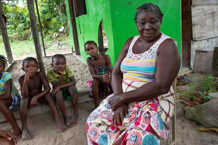 Emelien Adjako sits in her village of Kajapaati, Suriname, along the Suriname River, the damming of which caused her family to flee her flooding birth village when she was 5. When the creeks started to rise, she said, the elders “were praying to this tree, praying that the water wouldn’t come higher. So they were looking for their salvation with this tree, but it was the first thing to flood.” Image by Stephanie Strasburg. Suriname, 2017.<br />
