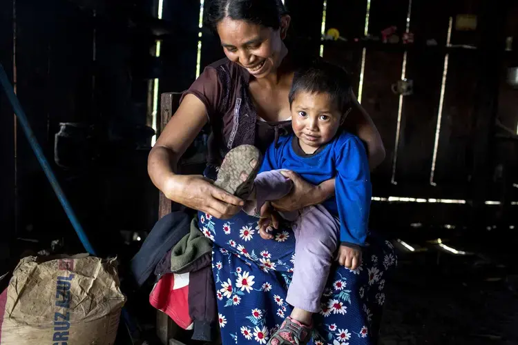 Catarina Domingo, dressing her son, lives in a house that is one of the few remaining in Yalambojoch that is made of wood and has a tin roof and a dirt floor. Image by Simone Dalmasso. Guatemala, 2019.<br />